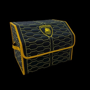 May include: Black storage box with yellow stitching and trim. The box features a geometric pattern and a yellow emblem. The box has a handle on the side and is set against a black background.