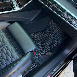 May include: Black car interior featuring leather seats with a quilted pattern and black floor mats with a white stitched design. The mats have a textured pattern and a red accent. The car's dashboard and door panel are visible.