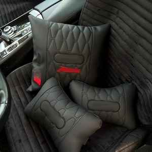 May include: Black car seat cushions and headrests. The cushions have a geometric quilted pattern with a rectangular accent. The headrests have a similar design. A red accent is visible on the larger cushion.