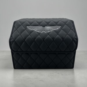 May include: Black quilted storage box with a diamond pattern. The box features a clear emblem on the front. The box is sitting on a gray surface. The box is designed for car storage.