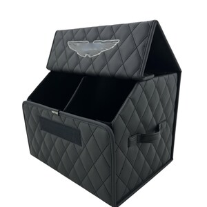 May include: Black car organizer with a quilted pattern and a clear emblem on the lid. The organizer has two compartments and a handle on the side. The word "Vortex" is printed on the front.