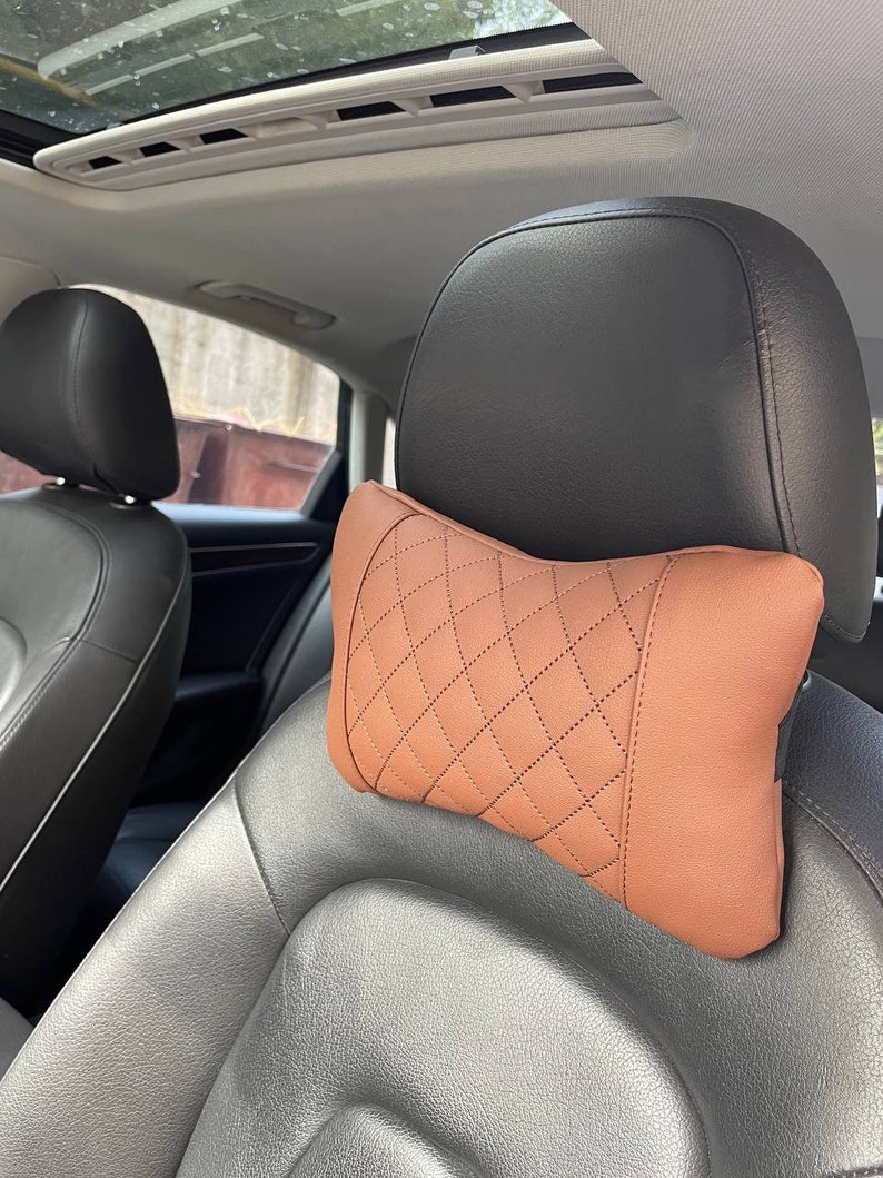 May include: A brown leather car headrest pillow with a diamond pattern. The pillow is designed to provide comfort and support for the head and neck while driving.