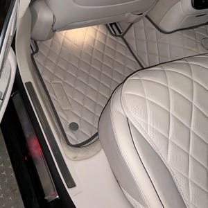 Puede incluir: Una alfombrilla de coche de color beige claro con un patrón de costuras en forma de diamante y ribete gris oscuro, instalada en el interior del vehículo. La alfombrilla cuenta con un parche rectangular texturizado y una fijación circular.