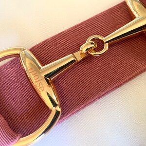 May include: A close-up of a gold-coloured metal belt buckle with the word "poise" engraved on it. The buckle is attached to a pink fabric belt.