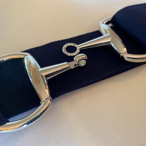 Peut inclure: Gros plan sur une sangle bleu marine avec des boucles en métal argenté. Les boucles sont gravées du mot "Louise".