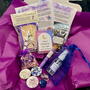 May include: A purple gift box filled with spiritual and magical items, including a pendulum, tea, candles, crystals, and a bottle of essential oil. The box is decorated with purple and blue patterned paper. The text on the cards reads "Spiritual Magickal Moments", "Divine Pendulum", "Opening Tea Brew", "Third Eye Tea", and "I am a channel for love, light and wisdom".