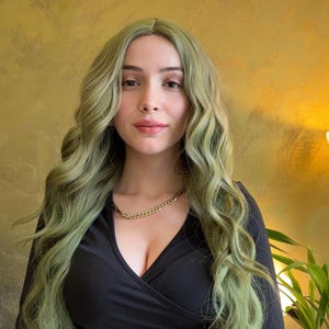 May include: A woman with long, wavy, light green hair and a gold chain necklace. She is wearing a black wrap-style top. The background is a textured gold color.