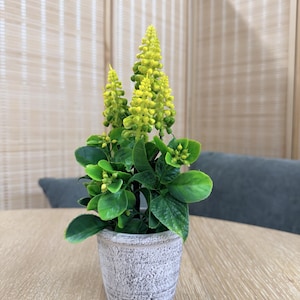 May include: A small artificial plant featuring three vibrant yellow, cone-shaped flower spikes rising above a cluster of deep green, textured leaves. The plant is housed in a cylindrical, weathered grey pot with a distressed, rustic finish.