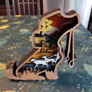 May include: A wooden dog silhouette with a layered design. The interior of the dog is cut out to reveal a scene of people walking dogs in a city park. The scene is painted in bright colours, including yellow, green, blue, and red.