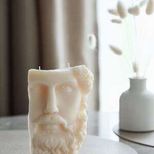 May include: A cream-coloured candle sculpted as a classical male bust with a beard and curly hair. The candle has two wicks and rests on a neutral surface. A white vase with dried flowers is in the background.