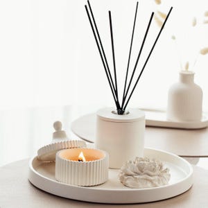 May include: A white tray displays a burning candle, a reed diffuser with black reeds, and a floral sculpture. The candle is in a ribbed container, and the diffuser is cylindrical. The items are arranged for home decor.