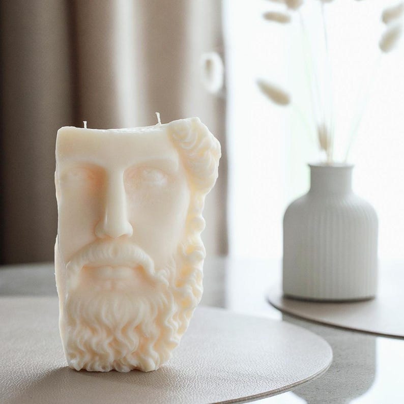 May include: A cream-coloured candle sculpted as a classical bust with a beard and curly hair. The candle has two wicks on top and is on a neutral surface. A white vase with dried flowers is in the background.