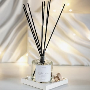 Op de afbeelding: Een heldere glazen diffuserfles met zwarte stokjes, op een wit dienblad. De fles heeft een label met de tekst "Golden Hour Home Fragrance". Een houten stopper ligt ernaast. De diffuser is een geurproduct voor in huis.