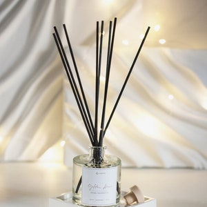 Op de afbeelding: Een heldere glazen fles met zwarte diffuserstokjes en een wit label met de tekst "Golden Hour Home Fragrance" op een witte dienblad.