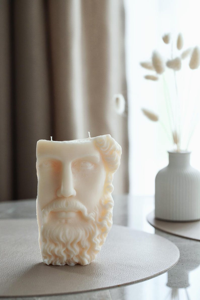 May include: A cream-coloured candle sculpted as a classical male bust with a beard and curly hair. The candle has two wicks and rests on a neutral surface. A white vase with dried flowers is in the background.