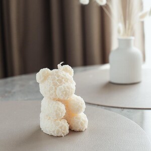 Op de afbeelding: Een witte teddybeerkaars gemaakt van kleine witte rozen. De kaars staat op een lichtbruin oppervlak.