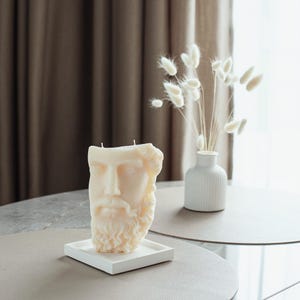 May include: A cream-coloured candle sculpted as a classical male bust with a beard, on a white square tray. The candle has two wicks. A small white vase with dried flowers is on a table in the background.