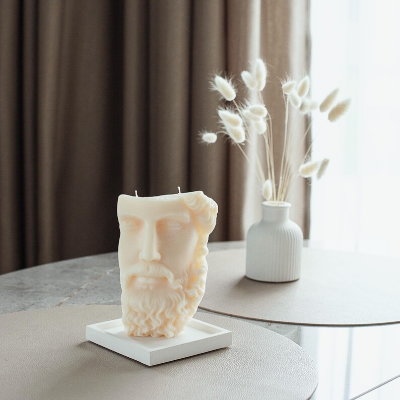 May include: A cream-coloured candle sculpted as a classical male bust with a beard, on a white square tray. The candle has two wicks. A small white vase with dried flowers is on a table in the background.