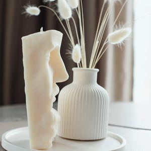 May include: A cream-coloured candle shaped like a face, next to a white vase with dried flowers. The candle is on a white tray. The vase has vertical ridges. The dried flowers are white and fluffy.
