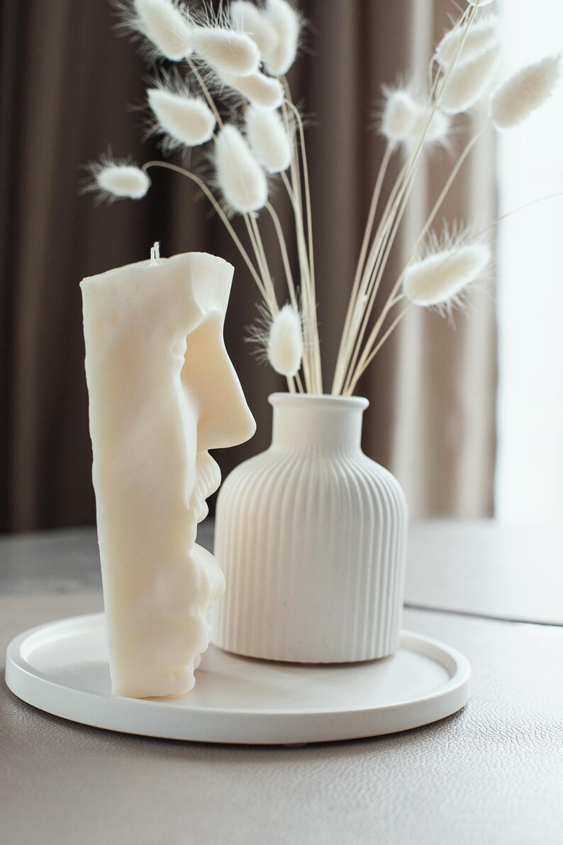 May include: A cream-coloured candle shaped like a face, next to a white vase with dried flowers. The candle is on a white tray. The vase has vertical ridges. The dried flowers are white and fluffy.