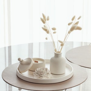 Puede incluir: Bandeja de cerámica blanca con un portavelas de cerámica blanca, un jarrón de cerámica blanca con hierba de la pampa blanca seca y una flor de cerámica blanca sobre una superficie marrón claro.