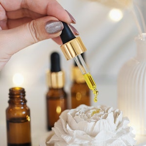 May include: A dropper dispenses yellow liquid onto a white, flower-shaped object. Amber glass bottles with gold-coloured caps are in the background. The scene suggests a skincare or aromatherapy product.