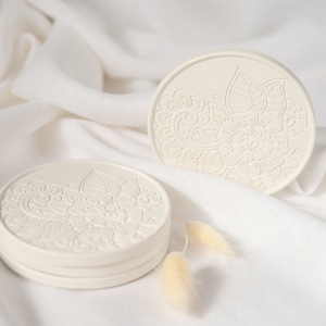 May include: Two white ceramic coasters with an intricate floral design. The coasters are stacked on top of each other and are sitting on a white fabric surface.