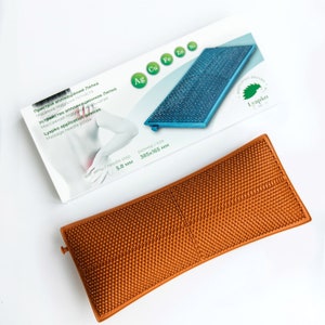 Pode incluir: Um travesseiro de agulha de massagem retangular marrom com uma superfície texturizada. O travesseiro é feito de plástico e tem uma forma curva. O travesseiro mede 385 mm por 165 mm e tem um passo de agulha de 5,8 mm. O travesseiro é rotulado como "Lyapko" e tem um logotipo de folha verde.