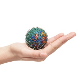Puede incluir: Una mano sostiene una pelota puntiaguda multicolor con un diseño verde, azul, rojo y amarillo. La pelota está cubierta de pequeños puntos afilados.