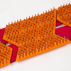 May include: Orange and red spiked traction device with a series of metal spikes on each section. The device is designed to be worn on shoes for added traction on slippery surfaces.
