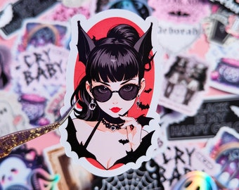May include: A sticker featuring a cartoon illustration of a woman with black hair, wearing a black choker, a red dress, and bat ears. She is wearing sunglasses and has a red background.