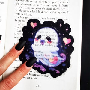 Może przedstawiać: Naklejka z duchem w kolorze czarnym i białym z galaktycznym wzorem w odcieniach fioletu, różu i błękitu. Naklejka ma białą obwódkę i zawiera serce z napisem "tro" w środku. Naklejka jest umieszczona na książce z tekstem po francusku.