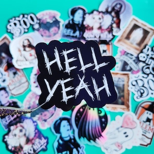 May include: A black and white sticker with the text "HELL YEAH" written in a bold, scratchy font. The sticker is surrounded by other colorful stickers with various designs, including ghosts, skeletons, and other Halloween-themed imagery.
