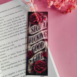 Pode incluir: Marcador de livros retangular com um design em estilo de tatuagem. Apresenta rosas vermelhas e o texto "SUCH A FUCKING GOOD GIRL" em uma faixa. O marcador está em um livro com texto.