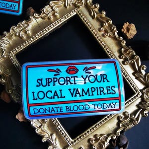 Może przedstawiać: Naklejka w kolorze niebieskim i czerwonym z tekstem "Support Your Local Vampires" i "Donate Blood Today". Naklejka przedstawia czerwony usta wampira z kłami i kapiącą krwią.