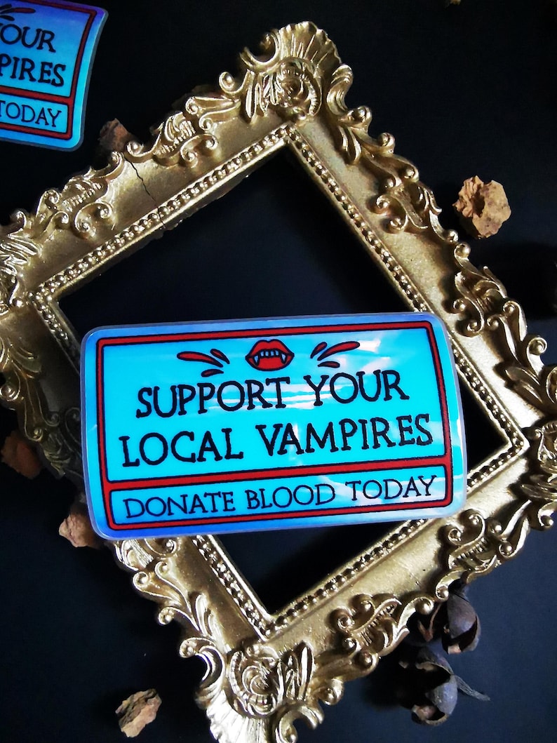 Może przedstawiać: Naklejka w kolorze niebieskim i czerwonym z tekstem "Support Your Local Vampires" i "Donate Blood Today". Naklejka przedstawia czerwony usta wampira z kłami i kapiącą krwią.