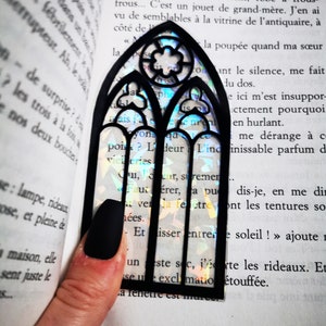 May include: A black bookmark shaped like a stained glass window with iridescent rainbow colors. The bookmark is placed on an open book with text.