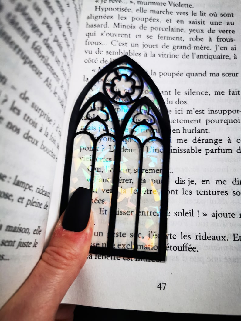 May include: A black bookmark shaped like a stained glass window with iridescent rainbow colors. The bookmark is placed on an open book with text.