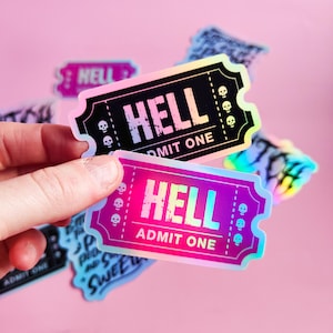 Może przedstawiać: Dwie holograficzne naklejki z motywem biletu. Naklejki są czarne i różowe ze słowem "HELL" i "ADMIT ONE". Małe ikony czaszki znajdują się na krawędziach. Inne naklejki są widoczne w tle.