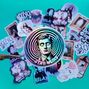 May include: A holographic sticker with a black and white illustration of a person with glasses and the text "Stay Weird". The sticker is surrounded by other stickers with various designs, including ghosts, skulls, and other spooky imagery.