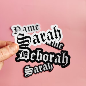 Op de afbeelding: Vijf witte stickers met zwarte letters. De stickers zijn in gotische letter en spellen de namen "Name", "Sarah", "Name", "Deborah" en "Sarah".