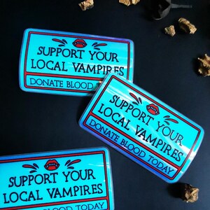 Może przedstawiać: Trzy niebieskie i czerwone naklejki z tekstem "Support Your Local Vampires" i "Donate Blood Today".