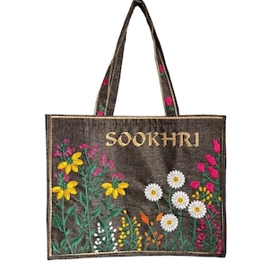 以下が含まれることがあります： ゴールドでSOOKHRIと刺繍されたダークブラウンのトートバッグ。バッグには、黄色、白、ピンクの花と緑の茎や葉を含むカラフルな花の刺繍が施されています。持ち手は本体と同じダークブラウンのものが2本付いています。