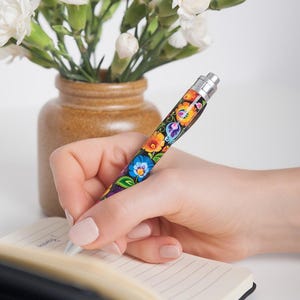 Könnte beinhalten: Ein einziehbarer Kugelschreiber mit einem lebendigen Blumenmuster aus orangefarbenen, blauen und grünen Blütenblättern auf schwarzem Grund. Der Stift hat einen silberfarbenen Metallclip sowie eine Spitze und liegt auf einem offenen Notizbuch.