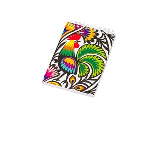 May include: A spiral-bound notebook featuring a vibrant, multi-coloured graphic illustration of a rooster. The design incorporates bold black, green, yellow, red, and blue patterns against a white background, showcasing stylised floral elements.