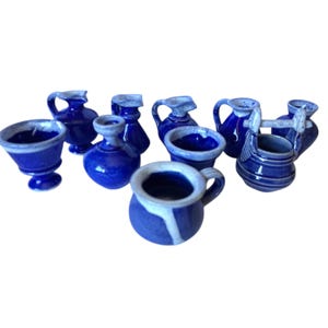 May include: A collection of miniature blue ceramic jugs, cups, and vases with white detailing. The assortment includes various shapes and sizes, with handles and spouts. The pieces are arranged on a white background.