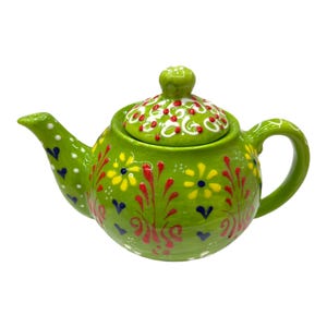 Può includere: Una teiera in ceramica verde con coperchio, manico e beccuccio. La teiera è decorata con disegni floreali gialli, rossi e bianchi e forme di cuore blu. Il coperchio ha un pomello sopra.