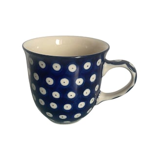 Op de afbeelding: Een keramische mok met een diepblauw glazuur en een herhalend patroon van witte stippen met donkerblauwe centra. De binnenkant van de mok is crèmekleurig en het handvat is versierd met hetzelfde blauw-witte stippenpatroon als de buitenkant.