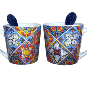 May include: Two white ceramic mugs with a colourful, geometric tile pattern in blue, red, yellow, and white. Each mug has a matching blue spoon resting inside. The mugs have a curved handle.