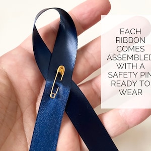 May include: A dark blue ribbon with a gold safety pin attached. The ribbon is tied in a loop and is ready to wear. The text "EACH RIBBON COMES ASSEMBLED WITH A SAFETY PIN READY TO WEAR" is visible in the background.